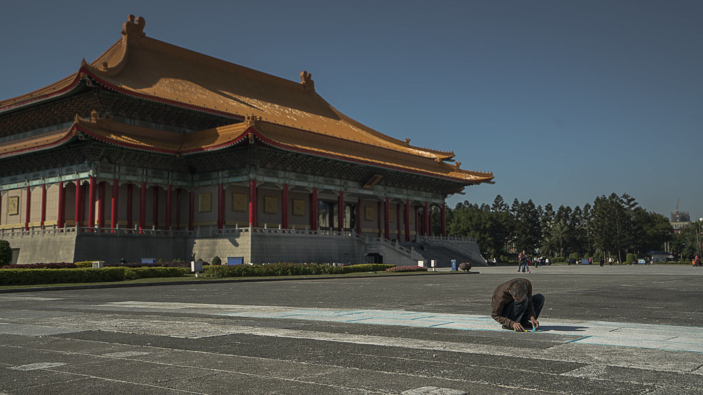 A man drawing with chalk on the plaza in front of a palace-roofed hall at Liberty Square