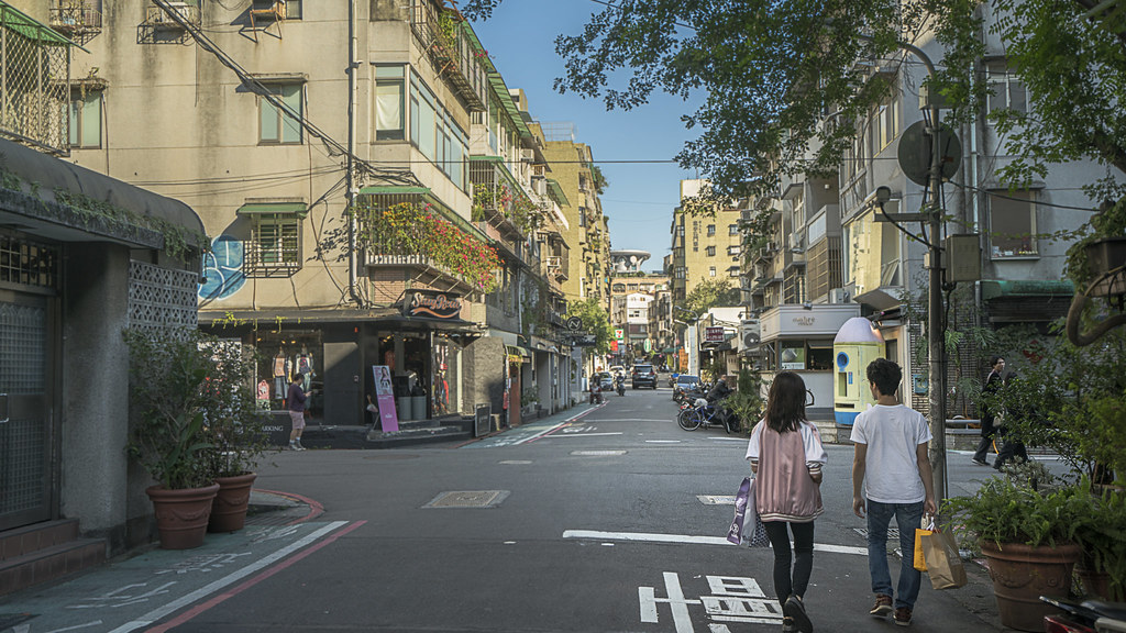 A quiet Taipei street corner with shophouses, balconies, and two people walking